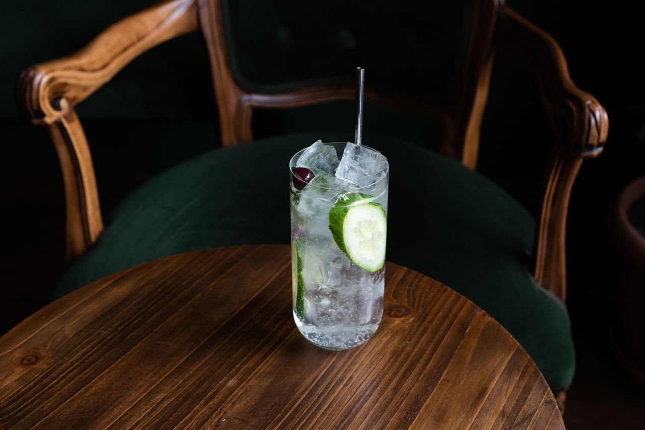 Highball glass filled with a gin and tonic, garnished with cucumber and a cherry, on a wooden table.