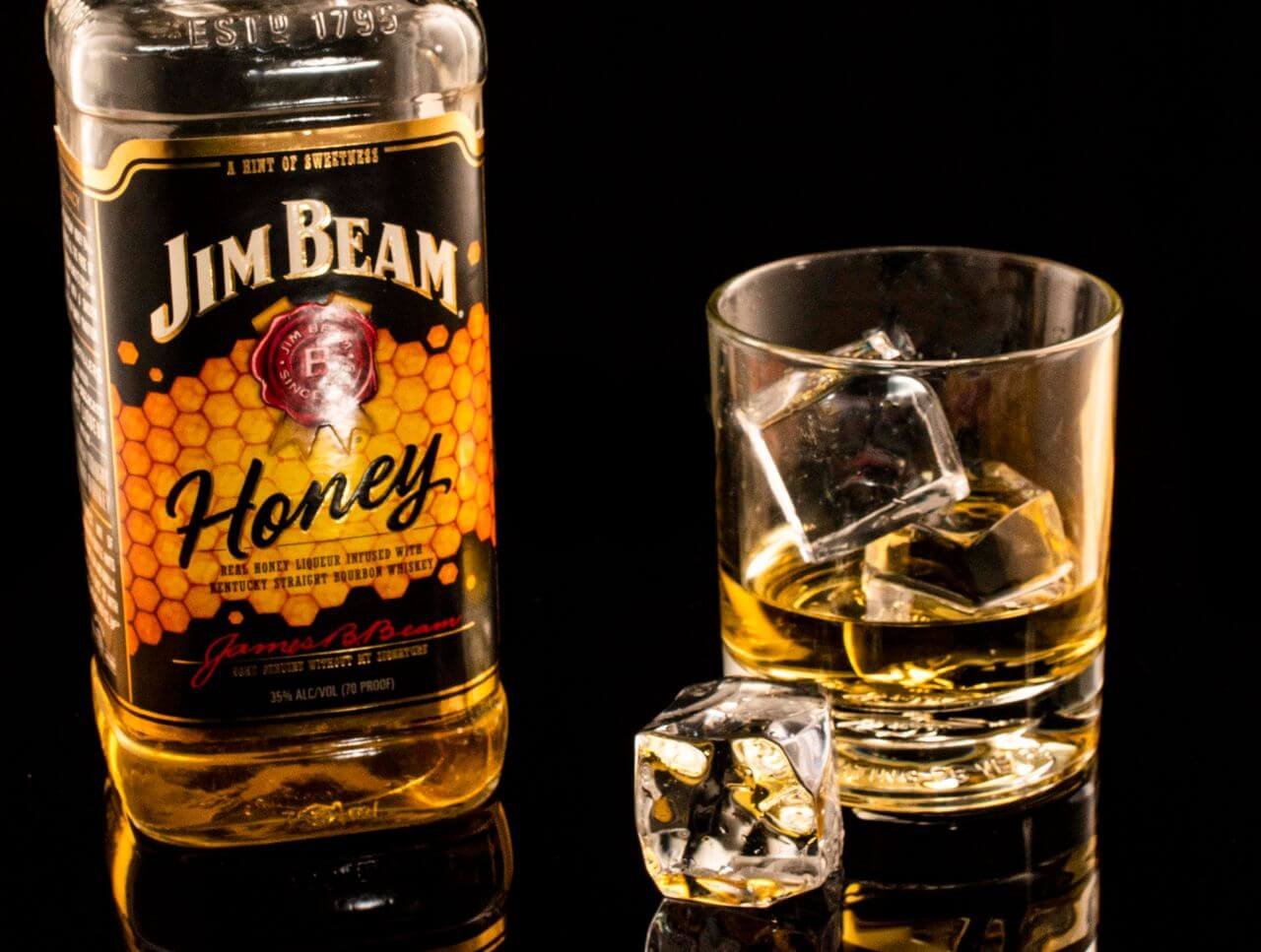 A bottle of honey whiskey beside a glass filled with whiskey and ice cubes on a reflective surface.
